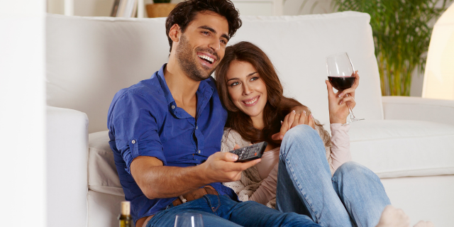 Casal sorrindo sentado no chão. Eles estão encostados em sofá branco. A mulher segura uma taça de vinho, enquanto o homem, ao lado dela, tem um controle remoto em mãos.