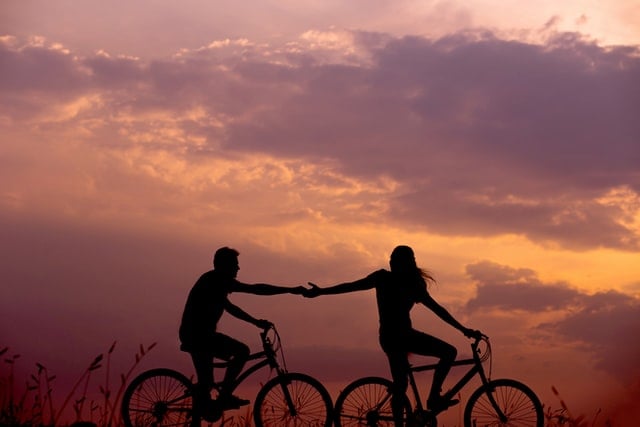 Homem e mulher pedalando bicicleta segurando as mãos durante pôr-do-sol