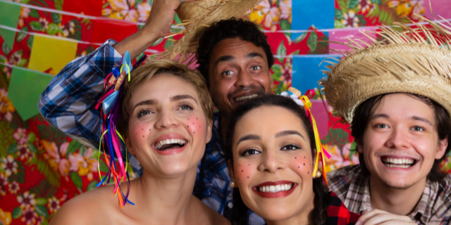 Dois homens e duas mulheres sorriem e comemoram festa junina. Eles estão tipicamente vestidos e o fundo é de tecido colorido e estampado.
