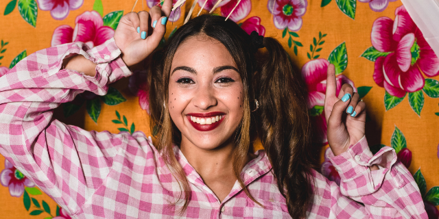 Mulher tipicamente vestida para festa junina sorri e ergue as mãos. O cenário é composto de um tecido amarelo estampado por flores.