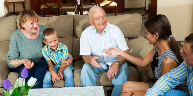 Neto sentado entre os dois avós em sofá. No sofá à frente, há um homem e uma mulher. Todos conversam e sorriem.