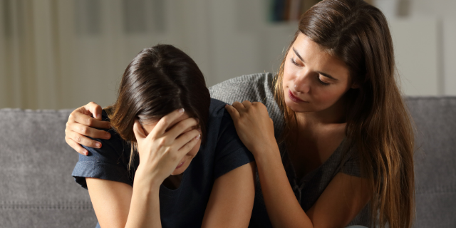 Duas mulheres sentadas em um sofá cinza. Uma abraça a outra e a consola, que está chorando com a mão sobre o rosto.