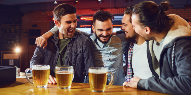 Quatro amigos homens em bar abraçados. Sobre o balcão há copos de cerveja.