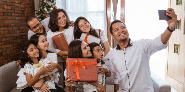 Homem tira selfie com a família. Eles estão sentados em um sofá e seguram presentes nas mãos.