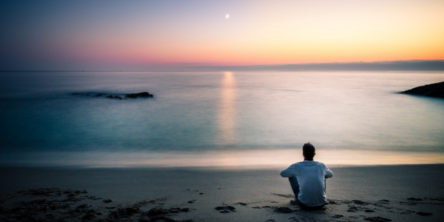 Homem sentado em areia observa ao pôr-do-sol. 