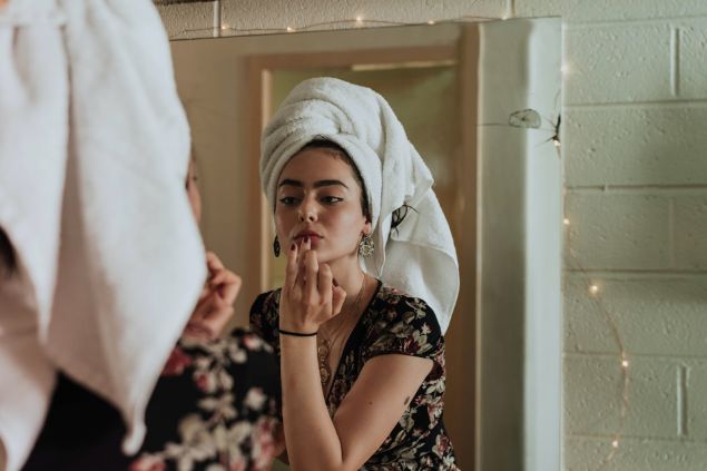 Mulher se maquiando em frente a um espelho, com uma toalha enrolada na cabeça.