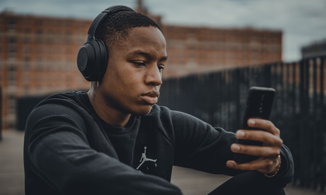 Homem negro sentado com fones no ouvido, enquanto segura celular.