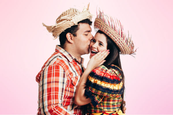 Homem e mulher com trajes de festa junina. O homem está beijando a bochecha da mulher.