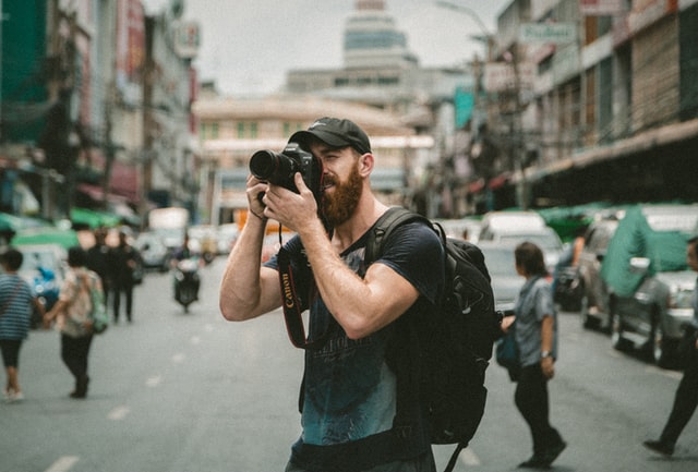 Homem branco segurando câmera fotográfica no meio da rua.