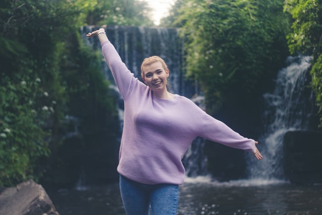 Mulher branca de cabelos presos e suéter lilás, de braços abertos, com cachoeira ao fundo.
