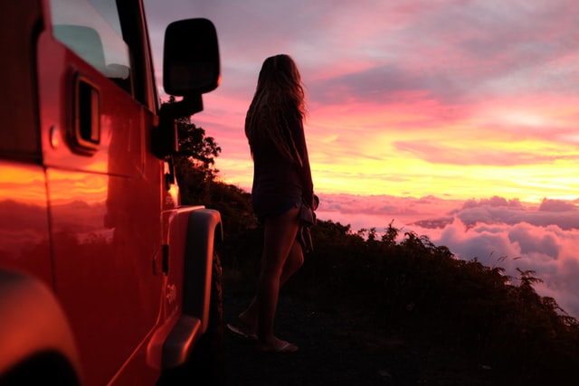 Mulher branca ao lado de carro vermelho enquanto olha o pôr-do-sol.