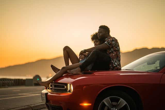 Homem e mulher negros abraçados sentados em cima de carro vermelho.