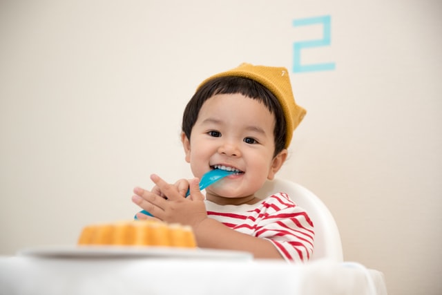 Bebê asiático com colher na boca e sorrindo.