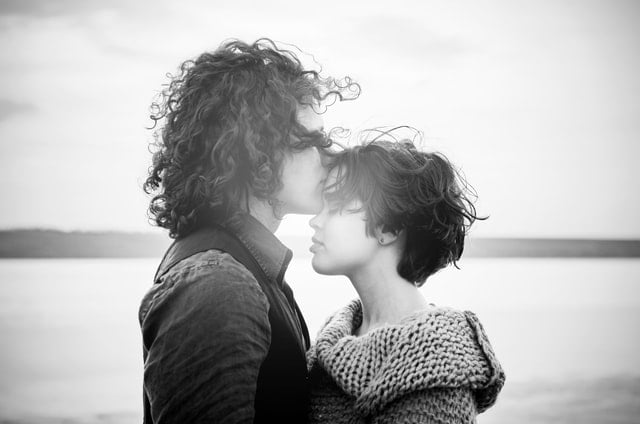 Homem branco beijando testa de mulher branca na praia.