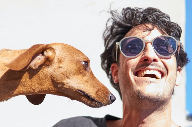 Homem branco de óculos escuros e expressão sorridente ao lado de um cachorro marrom.