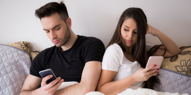 Um casal sentado em uma cama, ambos têm celulares em suas mãos.