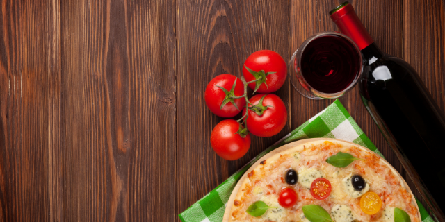 Pizza, tomates, garrafa e taças de vinho sobre superfície de madeira escura.