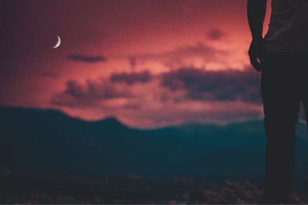 Silhueta parcial de um homem à direita e a Lua minguante ao longe, à esquerda, em um céu avermelhado.