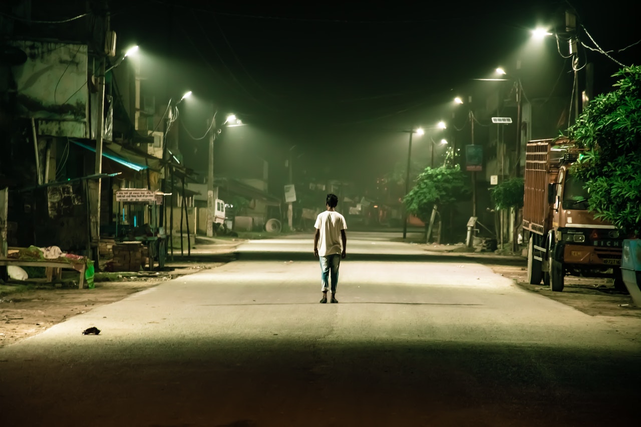 Homem andando sozinho em rua de cidade, deserta, durante a noite, apenas com as luzes dos postes.