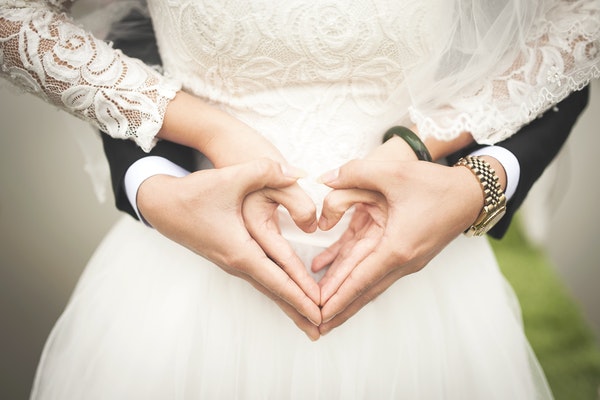 Mãos de casal formando coração com mulher de vestido de noiva e homem de terno