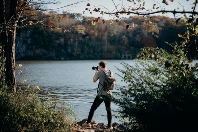 Homem branco fotografando um lago com árvores ao redor.