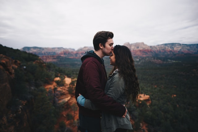 Homem branco beijando testa de mulher branca, com montanhas ao fundo.