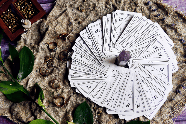 Várias cartas de Tarot viradas para baixo estão espalhadas sobre uma mesa, formando um círculo. No centro, há um cristal.  Ao lado, há plantas, flores e anéis.