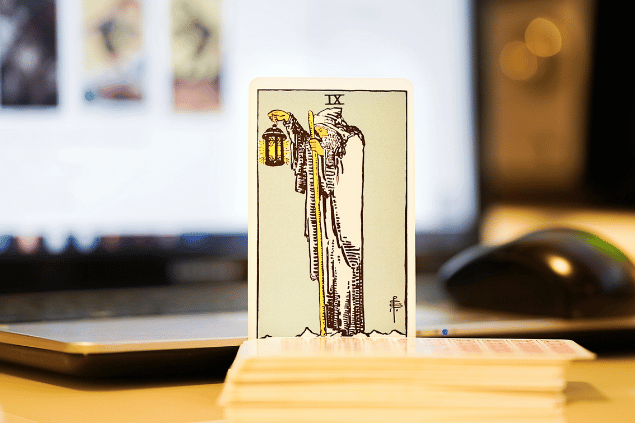 Na imagem, há a carta O Eremita do Tarot na posição normal e em pé. Ao fundo, há um notebook aberto.