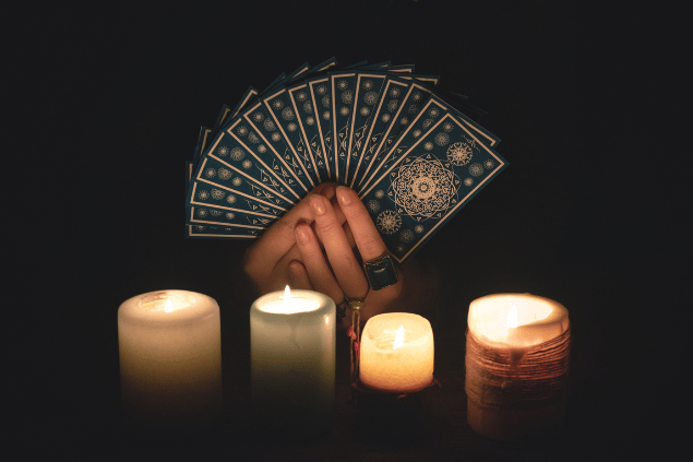 As mãos de uma taróloga segurando várias cartas de Tarot em sequência estão em destaque na imagem. Embaixo, há quatro velas acesas.