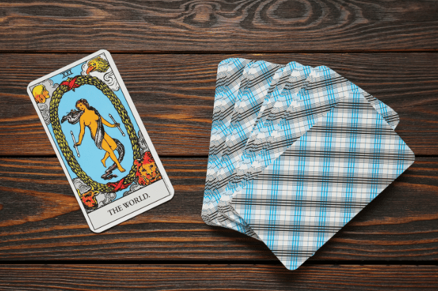 A carta O Mundo do Tarot está em destaque sobre uma mesa de madeira. Ao lado, há outras cartas de Tarot viradas para baixo.