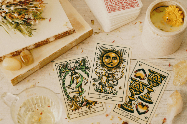 Três cartas de Tarot estão dispostas sobre uma mesa: O Mundo, O Sol e O Dois de Copas. Ao lado, há um baralho, flores, uma vela e cristais.