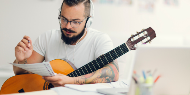 Homem com violão próximo ao corpo escreve em papéis que segura em uma das mãos. Ele está sentado e usa fones de ouvido.