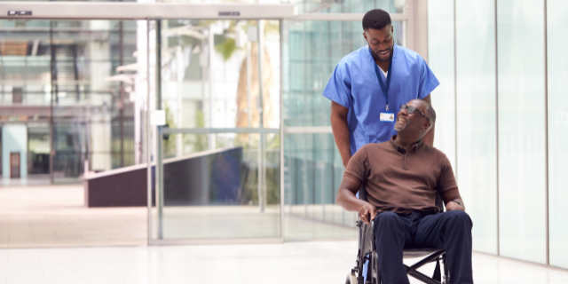 Enfermeiro empurra cadeira de rodas com um homem sentado sobre ela. Ambos sorriem. O cenário é hospitalar.