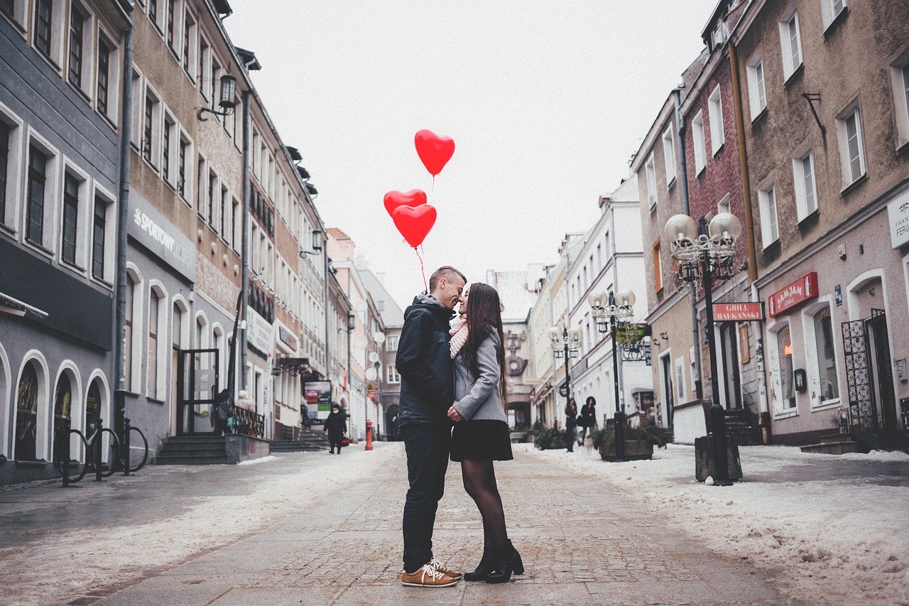 Homem e mulher frente a frente em uma rua, quase se beijando, segurando balões em formato de coração.