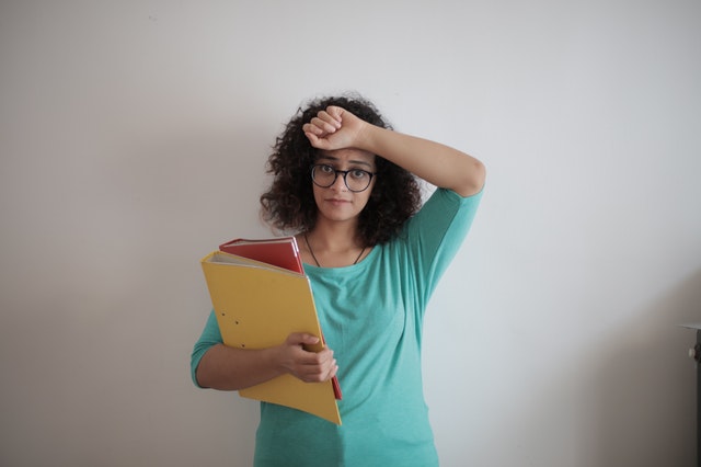 Menina negra de pele clara usando óculos, um braço segurando livros e outro levado à testa.