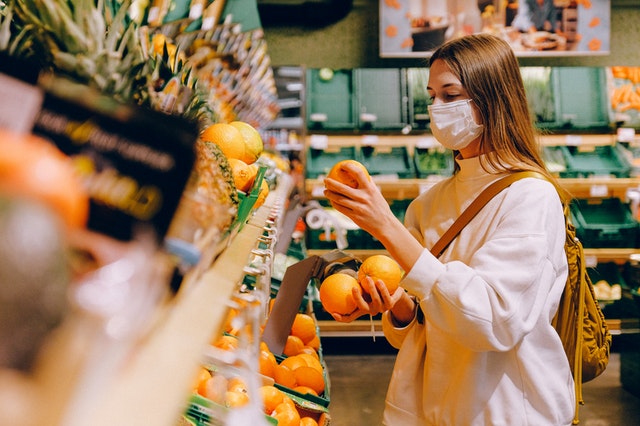 Mulher branca usando máscara num supermercado, enquanto segura laranjas.
