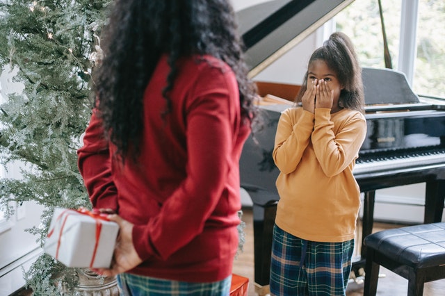 Uma menina à esquerda escondendo um presente enquanto uma menina à sua direita espera de maneira ansiosa com as mãos no rosto