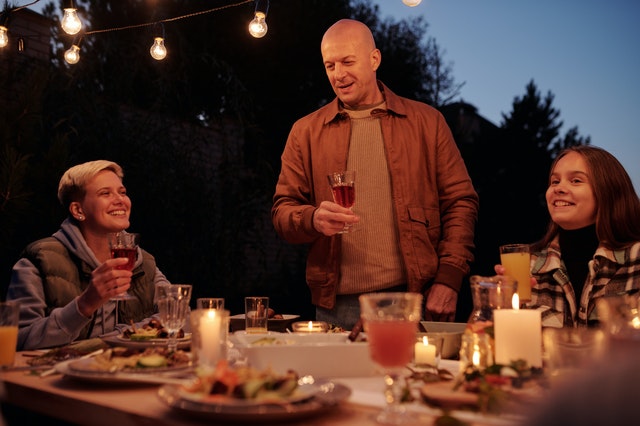 Homem em pé com taça de vinho na mão durante refeição em família ao redor de uma mesa farta de comida
