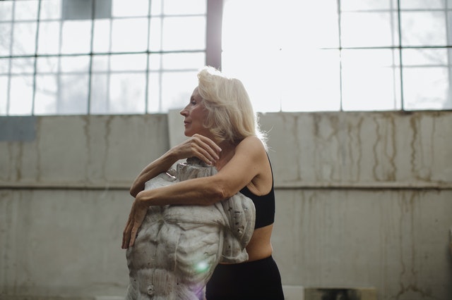 Mulher branca de cabelos brancos abraçadas à escultura.