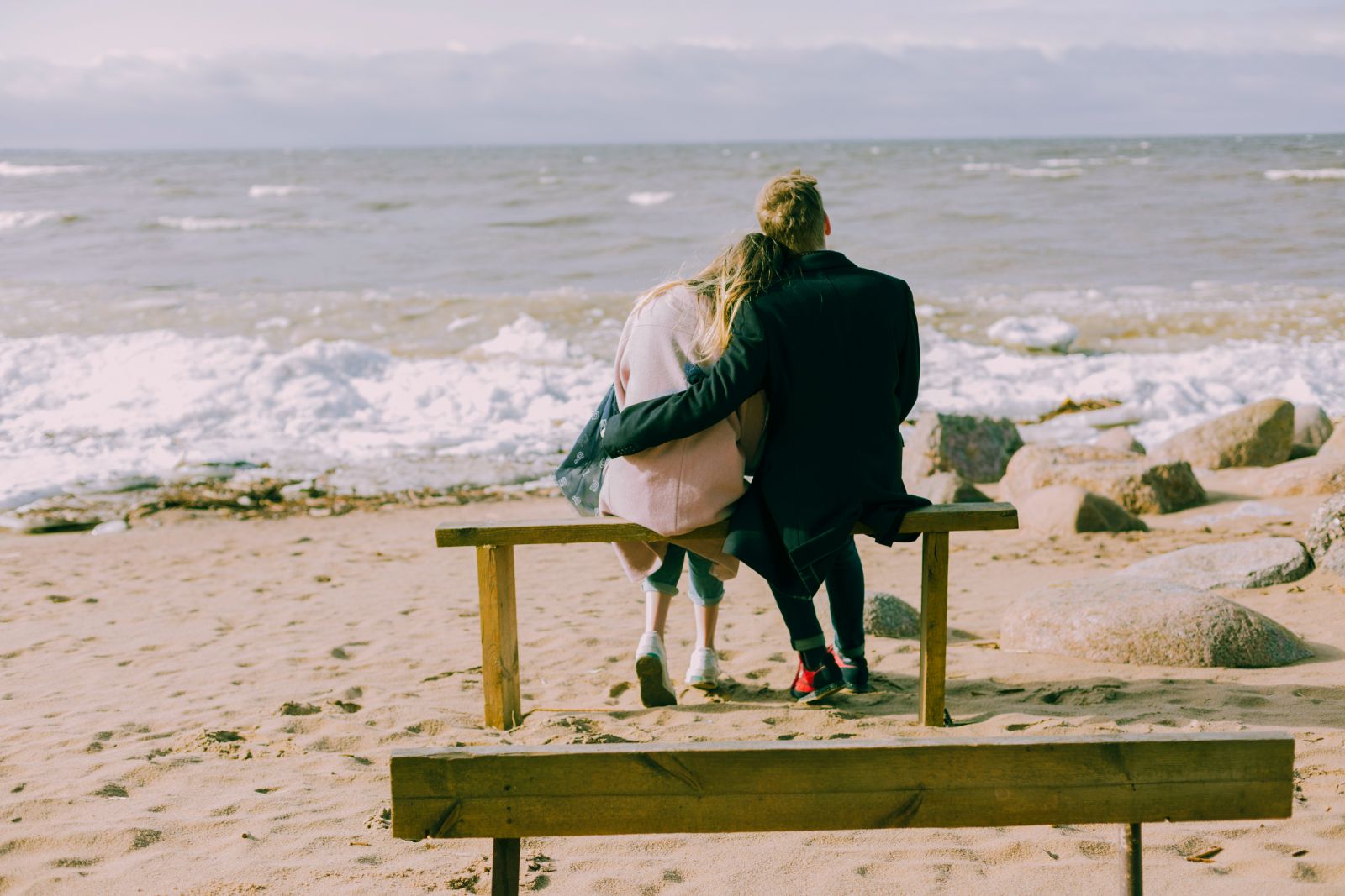 Casal sentado em um banco de madeira de frente para o mar da praia