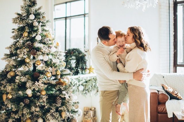 Pai e mãe beijando o filho no rosto ao lado de uma árvore de Natal