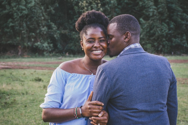 Homem e mulher negros abraçados, com homem dando beijo na bochecha da mulher.