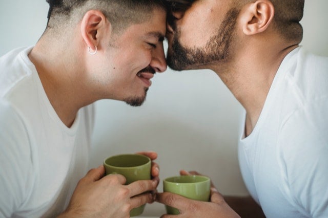 Homem beija a testa de seu companheiro enquanto os dois seguram xícaras de café