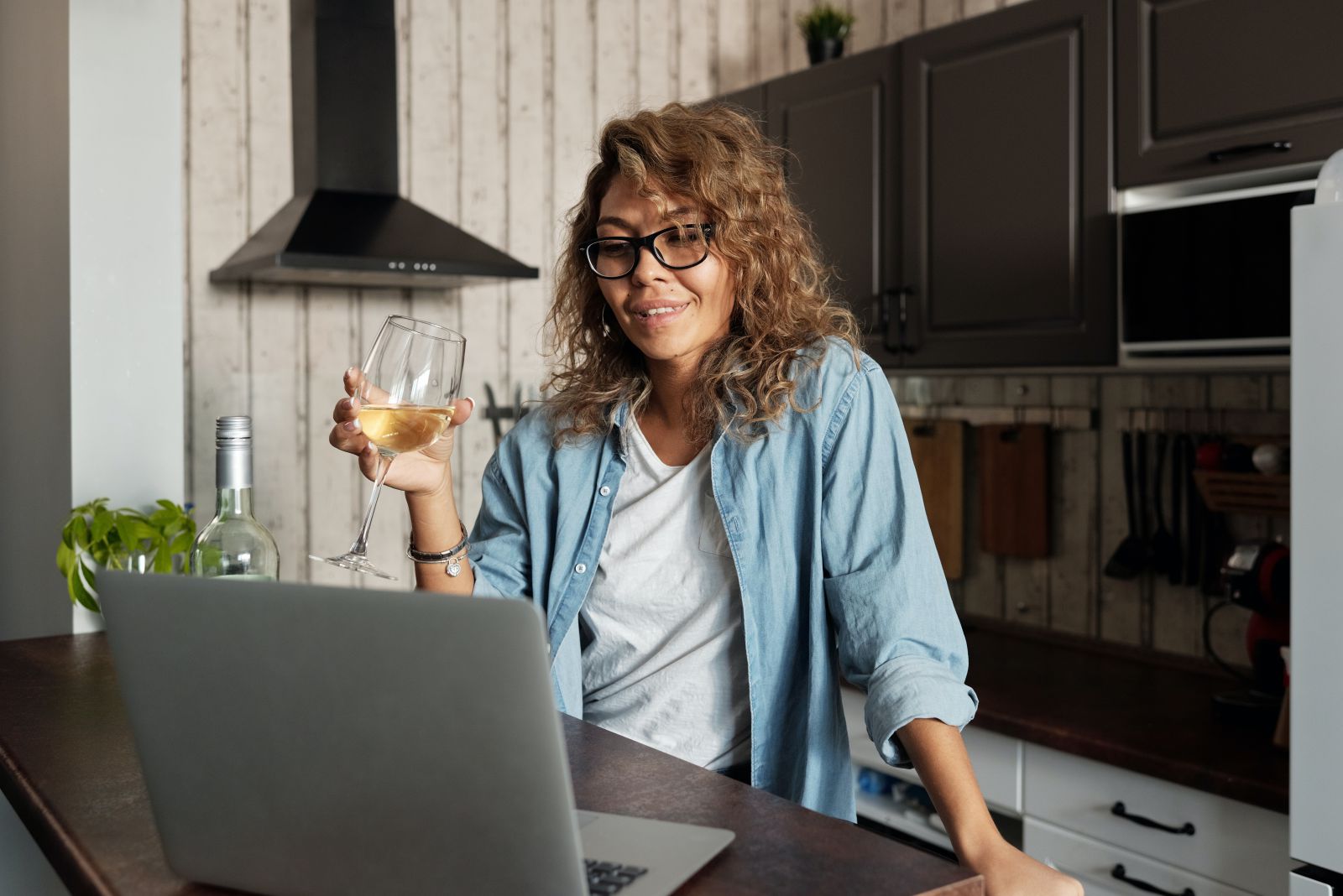 Mulher segurando taça de vinho em frente a um notebook na cozinha