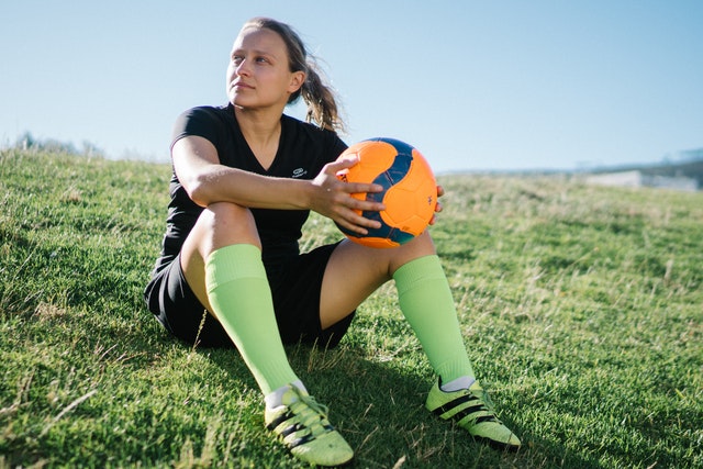 Jogadora branca sentada na grama com bola de futebol laranja nas mãos