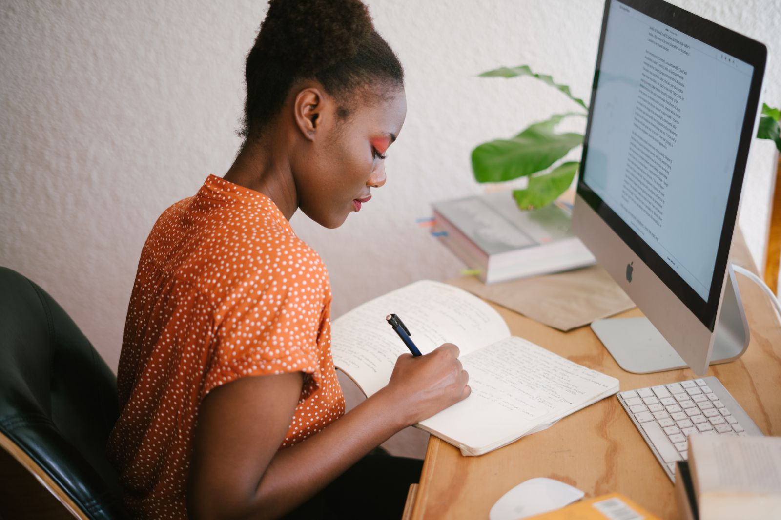 Mulher escrevendo em seu caderno em frente a tela de um computador
