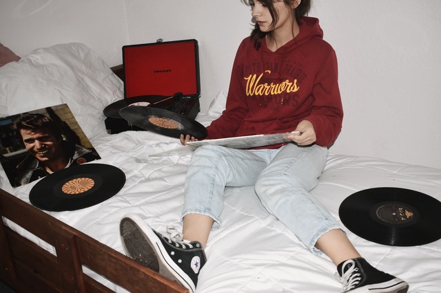 Menina branca sentada numa cama branca com discos e vitrola ao lado.