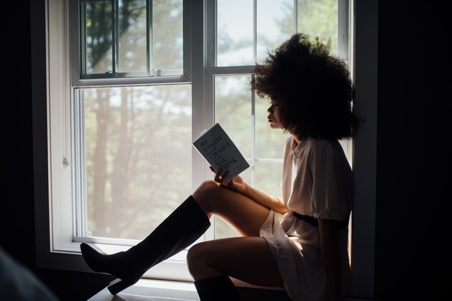 Mulher negra sentada na janela lendo livro.