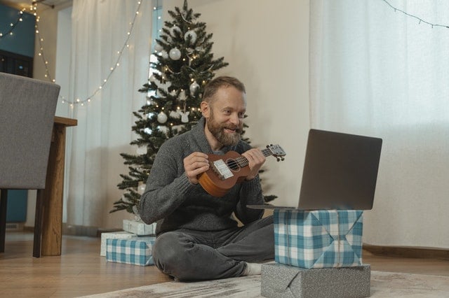 Homem sentado em um tapete mostrando um ukulele que ganhou de presente para alguém que está vendo ele pelo computador