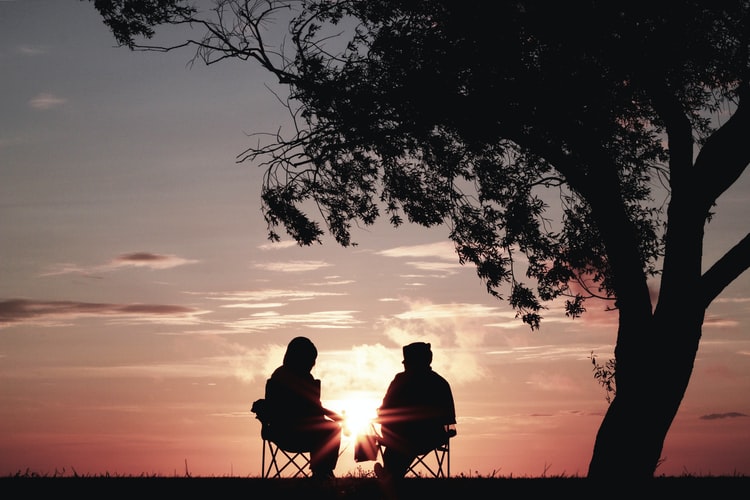 Duas pessoas sentadas assistindo um pôr do sol à esquerda de uma árvore